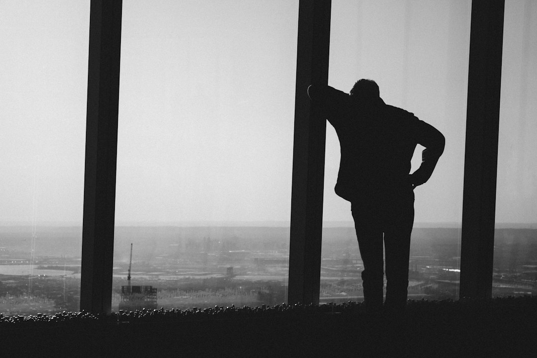 Un homme de dos, debout devant une grande baie vitrée, en noir et blanc